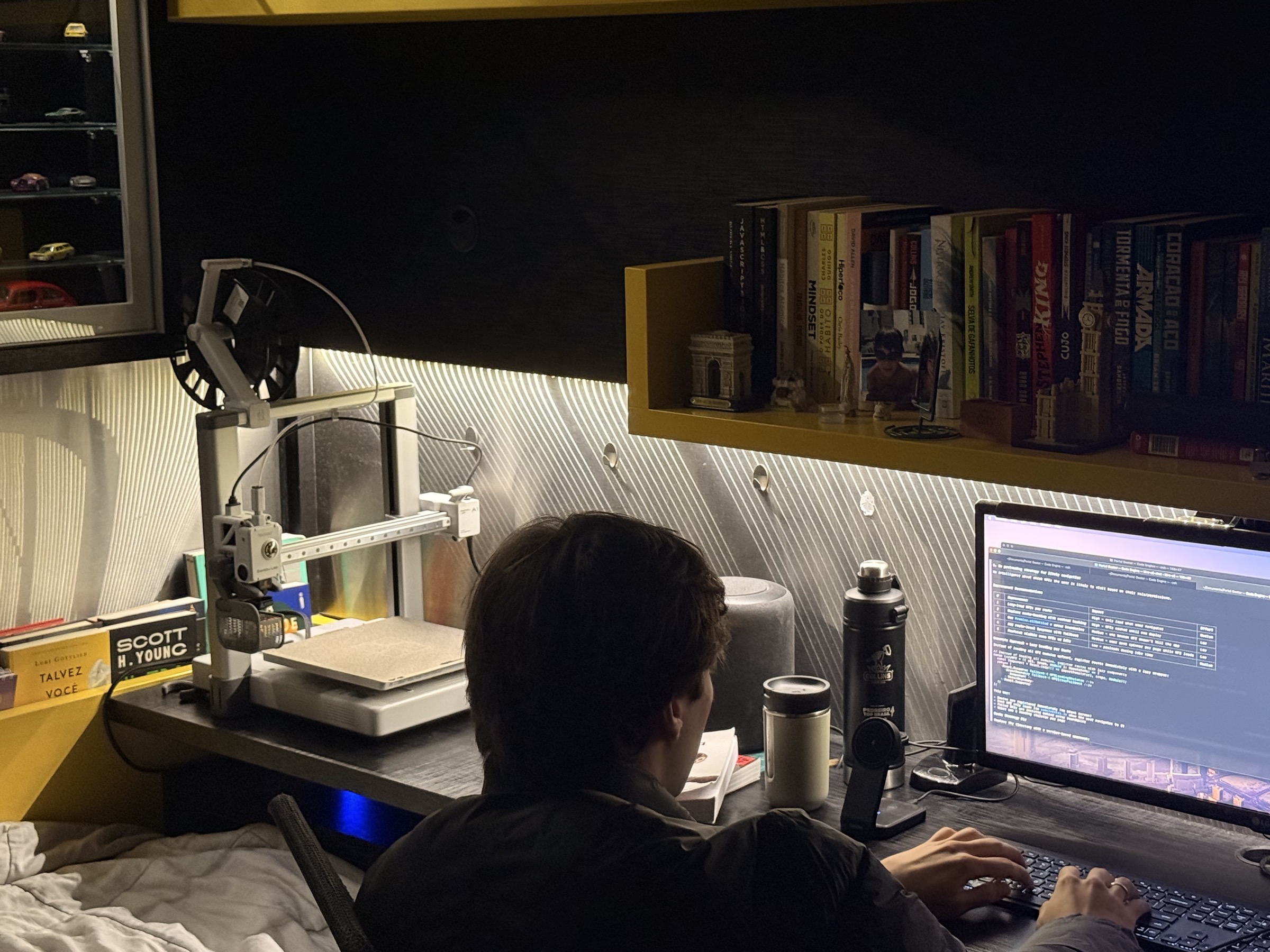 Bruno Fevereiro at his desk at night, coding beside a 3D printer and a shelf of books